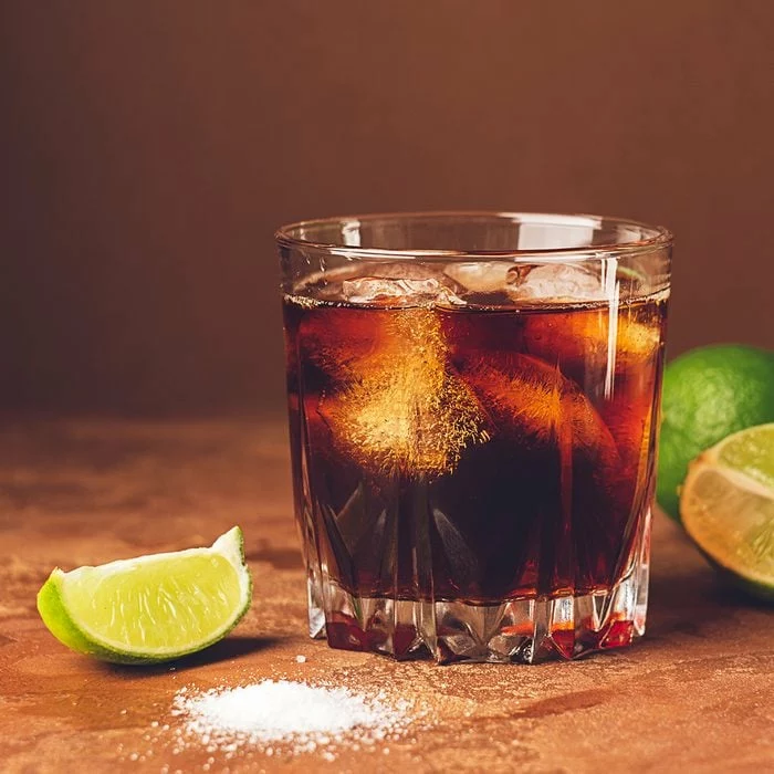 Cocktail froid avec de la glace et des morceaux de citron vert juteux dans un verre sur fond marron foncé.